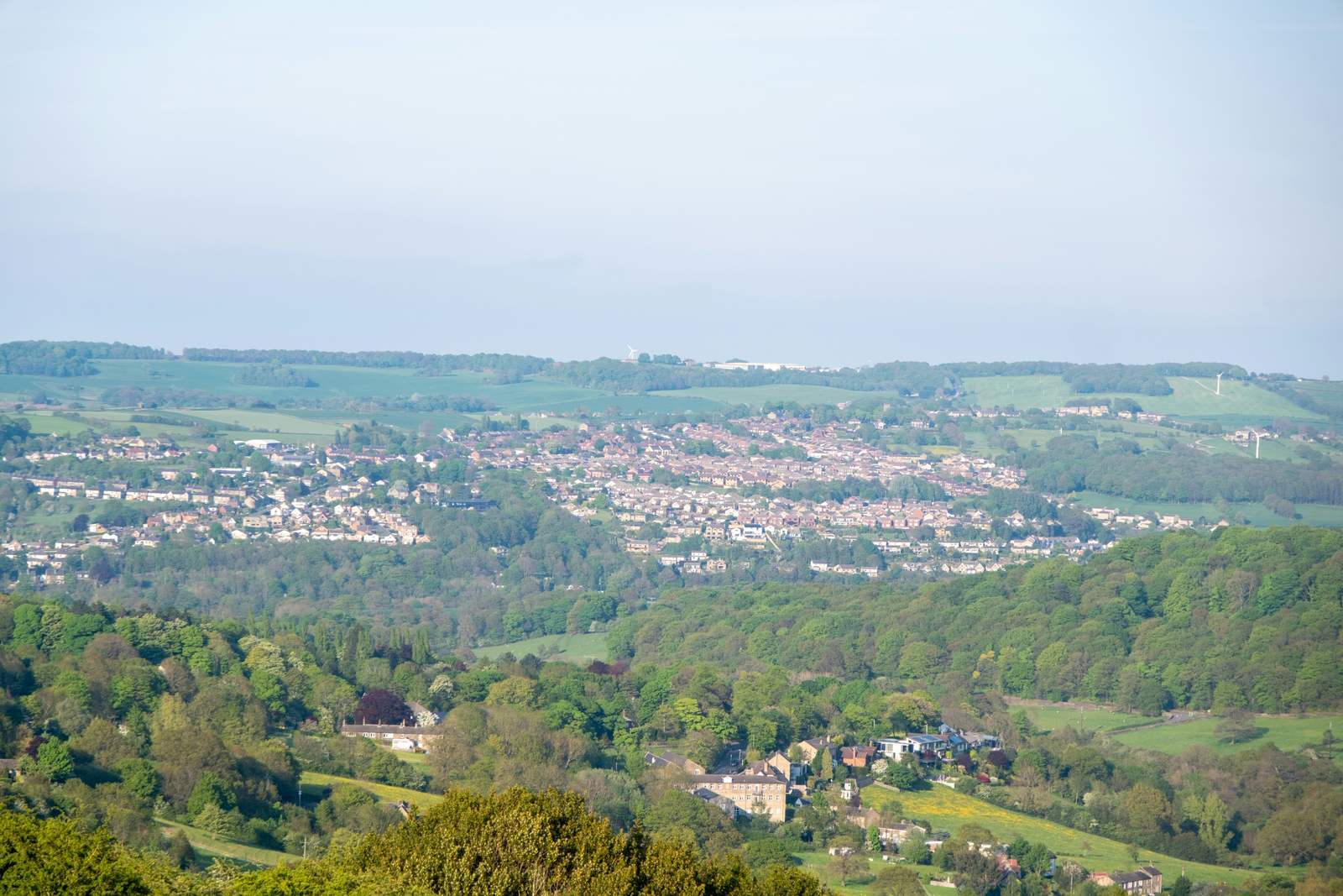 A town in a valley, surrounded by green fields and trees, with wind turbines on the distant hills